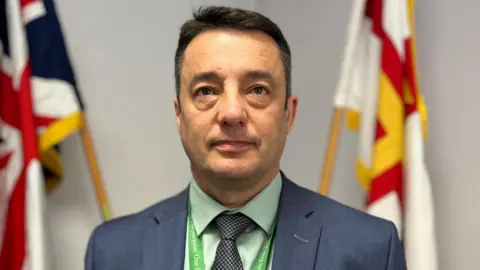 Boley Smillie - A man with a blue suit on a green shirt and a green lanyard standing in front of blurry Guernsey and British flags. 