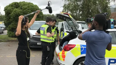 A camera operator and a sound recordist with a boom mic record two police officers standing by a police car