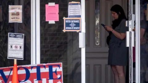 Reuters A woman stands looking at here phone at a polling station. A large sign reads "Vote Aqui, here" 