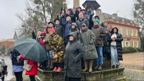 John Fairhall/BBC Scores of people surrounding a memorial wearing their raincoats. Some are standing on the ledge and wearing raincoats and others are holding umbrellas. They are trying to spot the King and Queen