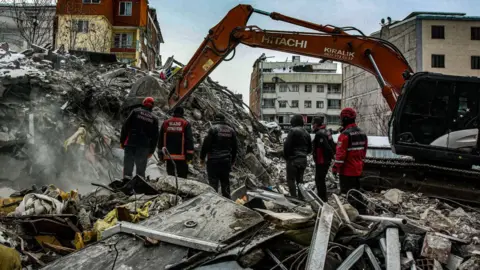 Getty Images Workers watch as a digger sorts through the rubble after the earthquake struck in Malatya, Turkey