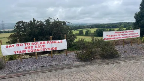 Two banners have been attached to wooden stakes at the top of a hill. The banner on the left reads "Say no to solar panels and save your countryside". The one on the right says "Maiden Law solar farm the size of 250 football pitches from Burnhope nature reserve to Lanchester".