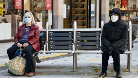 Getty Images Two people distancing on a bench in Cardiff outside shops, wearing masks