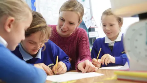 A female teacher sits with 3 school children aged about 10 years old wearing blue and white school uniforms .