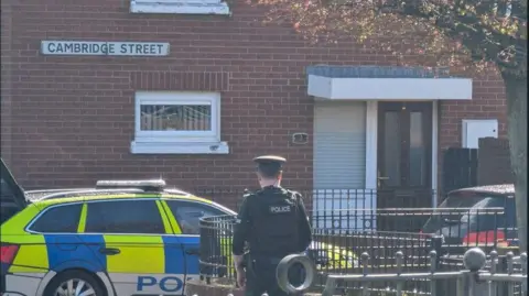 A police officer stands with his back to the camera - in front of him is a police car with battenberg markings. Behind the car is a brown-brick house with a small window, a brown PVC door and another window beside it. A street sign on the house reads "Cambridge Street".