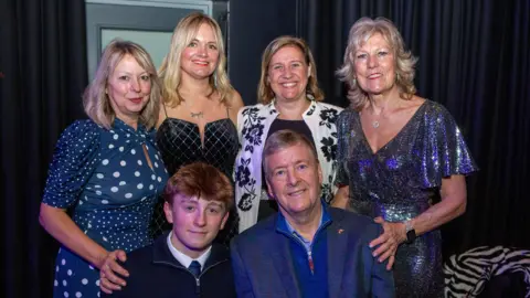 Four women stand up behind an older man and a teenage boy, both of whom are sat down. Everyone in the photograph is wearing blue and stood in front of a curtain and door at an awards ceremony.