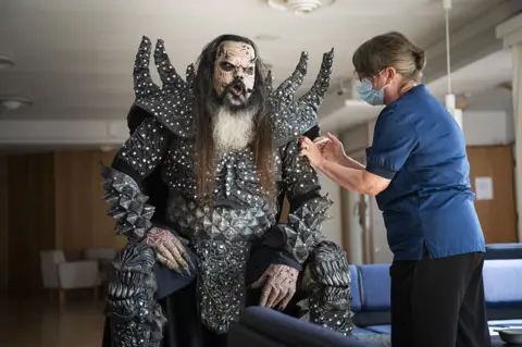 Jouni Porsanger / AFP Mr Lordi, aka Tomi Petteri Putaansuu, of the Finnish hard rock band Lordi, gets the second jab of his Covid-19 vaccination from a nurse in Rovaniemi, Finland, while wearing his rock band costume depicting him as a devil