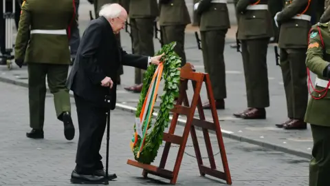 PA Media Michael D Higgins laying a green wreath with a ribbon in the colours of the Irish tricolour on a wooden stand. He is holding the wreath in one hand and a walking stick in the other. In the background a line soldiers in green military dress.