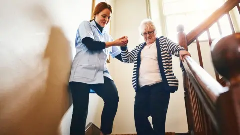 Dean Mitchell Getty Images Care worker helps elderly client down stairs