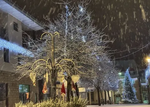 BBC Weather Watchers / Lyz Scarr Snow falling in Penicuik, Scotland during a cold snap in the UK. A tree can be seen in the foreground, with its branches covered in snow. There is a Christmas tree and decorations also visible in the picture, with fairy lights being hung from surrounding buildings