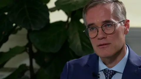 Chris Woodroofe with grey hair and black glasses wearing a blue suit and tie. He is sitting on a leather sofa with a large plant to his left. He looks impassively.