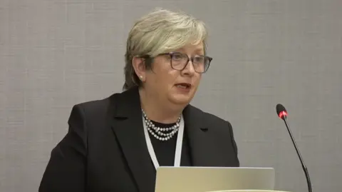 Eljamel Inquiry Joanna Cherry KC, a woman in her late 50s with short blonde hair and glasses speaks into a microphone. She is wearing a black jacket with pearls.