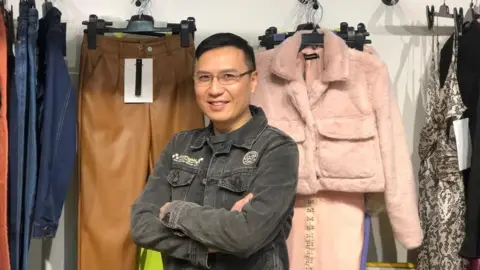 JNB Clothing Johnny Tseng in front of fashion pieces he sells to UK labels including a pink furry coat and camel trousers