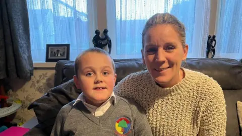 Oliver Conopo/BBC Young boy in grey sweater sitting next to woman with short light-coloured hair in a cream-coloured jumper