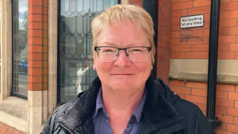 BBC/Amanda White A woman with short cropped sandy hair and glasses, she smiles at the camera on a sunny day outside an old brick building.