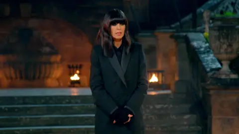 BBC Claudia Winkleman standing before the traitors up the palace steps with a metal basket of fire behind her