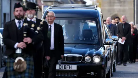 Alex Salmond's funeral procession heading from the church, led by a bagpiper 