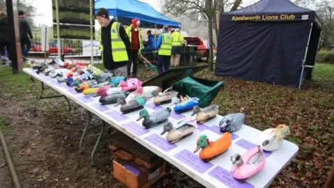 Kenilworth Lions Club A man in a black hat and high-vis jacket is standing behind a white table filled with two rows of decorated plastic ducks