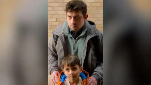 A man in a grey jacket stands with his hands on a boy's shoulders. He looks concerned.