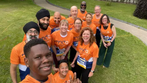 A group of 13 people are standing on a grassy area taking a selfie. Some are wearing orange, white and blue running bibs and some are wearing T-shirts of the same colour. They are wearing running numbers 