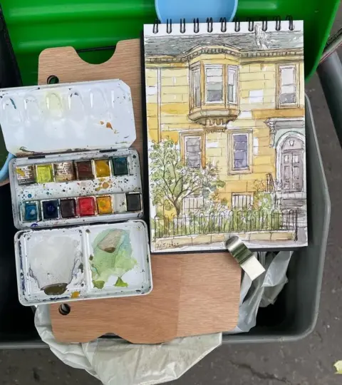 Allan Richardson A photograph of a watercolour paint palette and sketchbook with a painting of a tenement flat in Glasgow's west end resting on the top of a street sweeping cart.