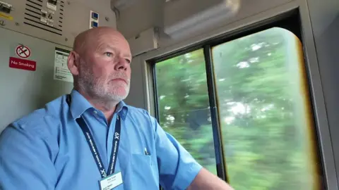 Peter Cook is seen in his cab while driving a train. He is bald with a short, white beard and wears an ID lanyard over a blue, short-sleeved shirt.