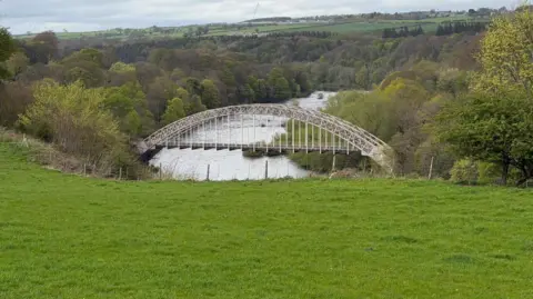 Lawrence O'Donnell A single span bridge sits over a wide river at the bottom of a green field. Its banks behind are wooded and in the far distance more fields.