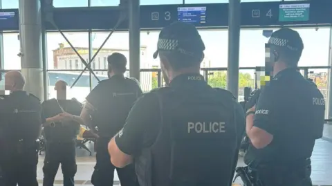 The picture shows from behind police officers wearing dark green uniforms, baseball caps and flak jackets. In from of them are three members of a Home Office enforcement team, also wearing dark uniforms. The picture is taken inside a bus station