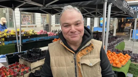 A man in a beige jacket smiling for the camera. Several varieties of fruit are stacked up in the background at a colourful fruit staff.