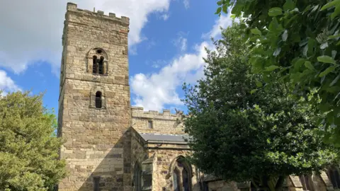 The crenelated tower of St Cuthbert's Church emerging over a tree. The roof of the main body of the church can also be seen.