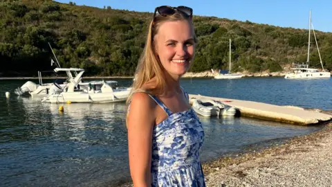 Supplied Anna Barker is pictured in blue dress in front of a river with boats in the background. She has long blond hair and sunglasses resting on the top of her head