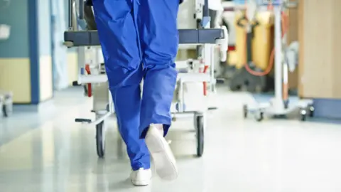 The legs of a person wearing blue medical scrubs and pushing a hospital trolley