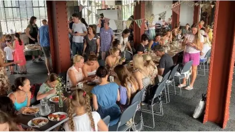 BBC Ukrainian refugees, adults and children, eating together on a long table stretching the length of a large room at The Pattern Church in Swindon