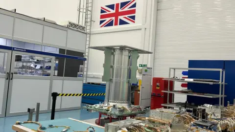 Components of a spacecraft assembly are laid out in an SSTL room, with wiring and metal structures on workbenches and a large cylindrical section standing upright beneath a Union Jack on the wall.