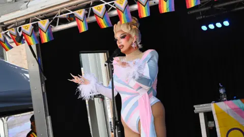 Haydn Brown A drag queen with a blonde wig tied back into a large bun. She has heavy make-up on with bright pink cheeks and dark eyes. She is standing on stage talking into a microphone while gesticulating with her hands while wearing a white, blue and pink leotard. The stage is decorated with small Pride flags.