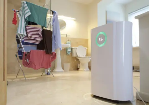 Getty Images A dehumidifier runs next to a pile of washing which is drying on a clothes rack in a large bathroom. The dehumidifier display reads 59%.