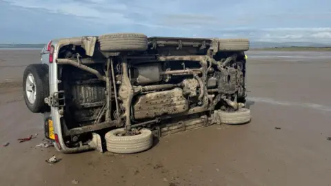 A car is flipped on its side. You can see the underside covered in sand. There is sand on the ground and the ocean in the background. It is a sunny day. 