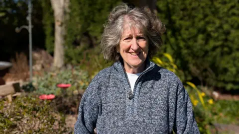A woman stands outside on a sunny day. She has curly grey hair and she is wearing a zip-up grey and blue jumper. Trees and flowers can be seen behind her.