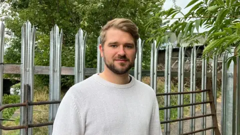 Ollie Conopo/BBC Man with light brown hair and short beard stands in front of closed-off footpath