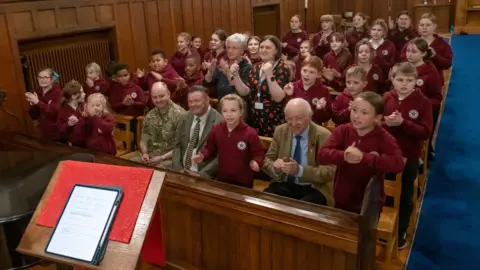 North Yorkshire Council Children sit singing in the church pews, encouraged by two men and two women sitting between them.