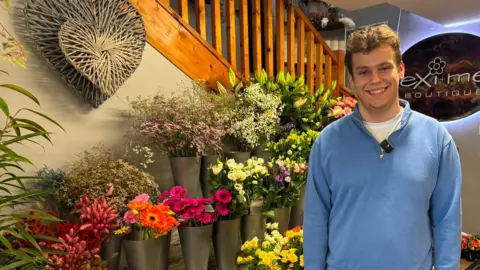 BBC Jamie is wearing a blue quarter zip sweater. He is smiling and behind him is an array of flowers and plants. 