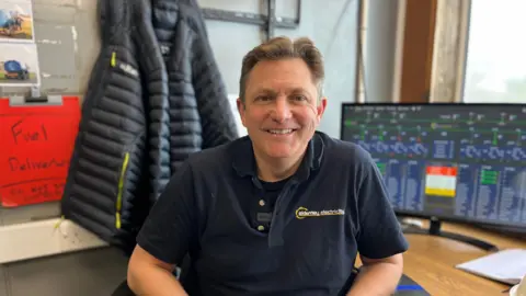 Chris MacGregor looking at the camera, sat in front of his desk which has a monitor on it.