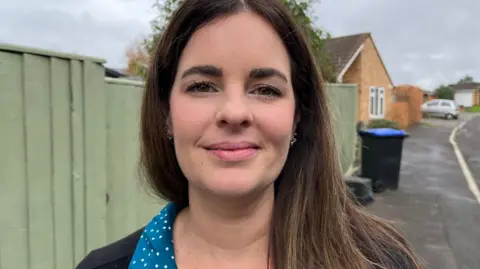 BBC Joeanne looking at the camera smiling. She has long brown hair and is wearing a blue shirt with white spots and a black top. Behind her is a light green fence. 