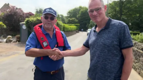 Two men shake hands on a rural road. The man on the left is wearing a blue baseball cap, sunglasses and red life jacket, on top of a blue polo shirt. The man on the right is wearing glasses and a navy polo shirt.