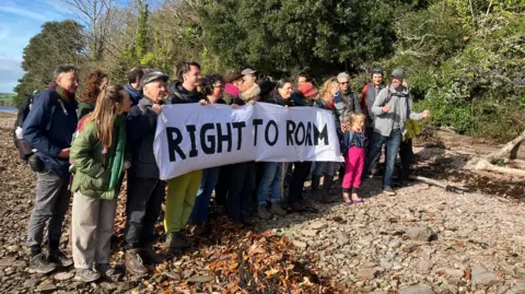 A group of people stood together holding a banner which says RIGHT TO ROAM. They are standing on the banks of a river and are wearing backpacks and coats.