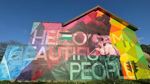 BEN SCHOFIELD/BBC A brightly coloured mural on the gable end of a house. It includes a rainbow of colours and depicts a man in a black knitted hat leaning over an hugging another person. The words "hello my beautiful people' are written over the top of the picture, blending with the colours. A small Palestinian flag, not part of the original artwork, has also been attached to the wall, over the to Ls in 'hello'. The sky is deep blue and cloudless, on a bright sunny day.  