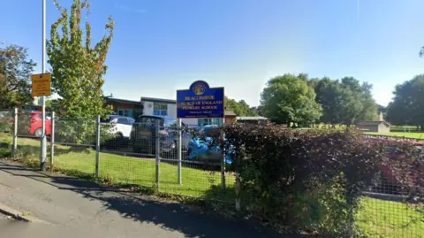 Outside Beaconside C of E Primary School in Hutton Hill, Penrith. There is a blue school sign with a crest and yellow writing, in front of it a wire fence and trees. The school buildings, which have sloping roofs are partially visible in the background.