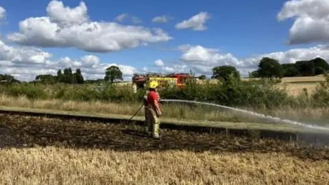 Avon Fire and Rescue Service A fire officer with a hose putting water on a scorched field with a fire engine in the backgorund