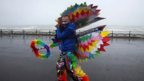 Reuters A man in a rainbow outfit battles the wind on Brighton seafront in front of choppy waves