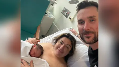 Claire Barlow Selfie of Claire and her husband with baby Osian in hospital. Claire is lying down on a bed in a hospital room smiling whilst holding her baby. Claire's husband is taking the selfie and he is also smiling.